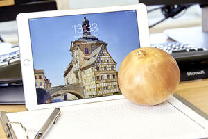 Bild vergrößern: Auf einem Tisch steht ein Tablet mit der Ansicht des Rathauses an der Oberen Brück, davor links ein Stift sowie rechts eine Zwiebel in Andeutung des Begriffs Zwiebeltreter.