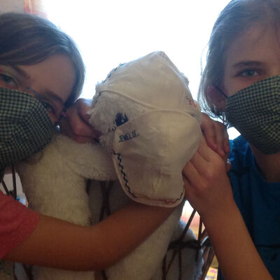 Bild vergrößern: Auf dem Bild sieht man zwei Schwestern, die einem Kuscheltier auch Mund-Nasen-Masken aufgesetzt haben.